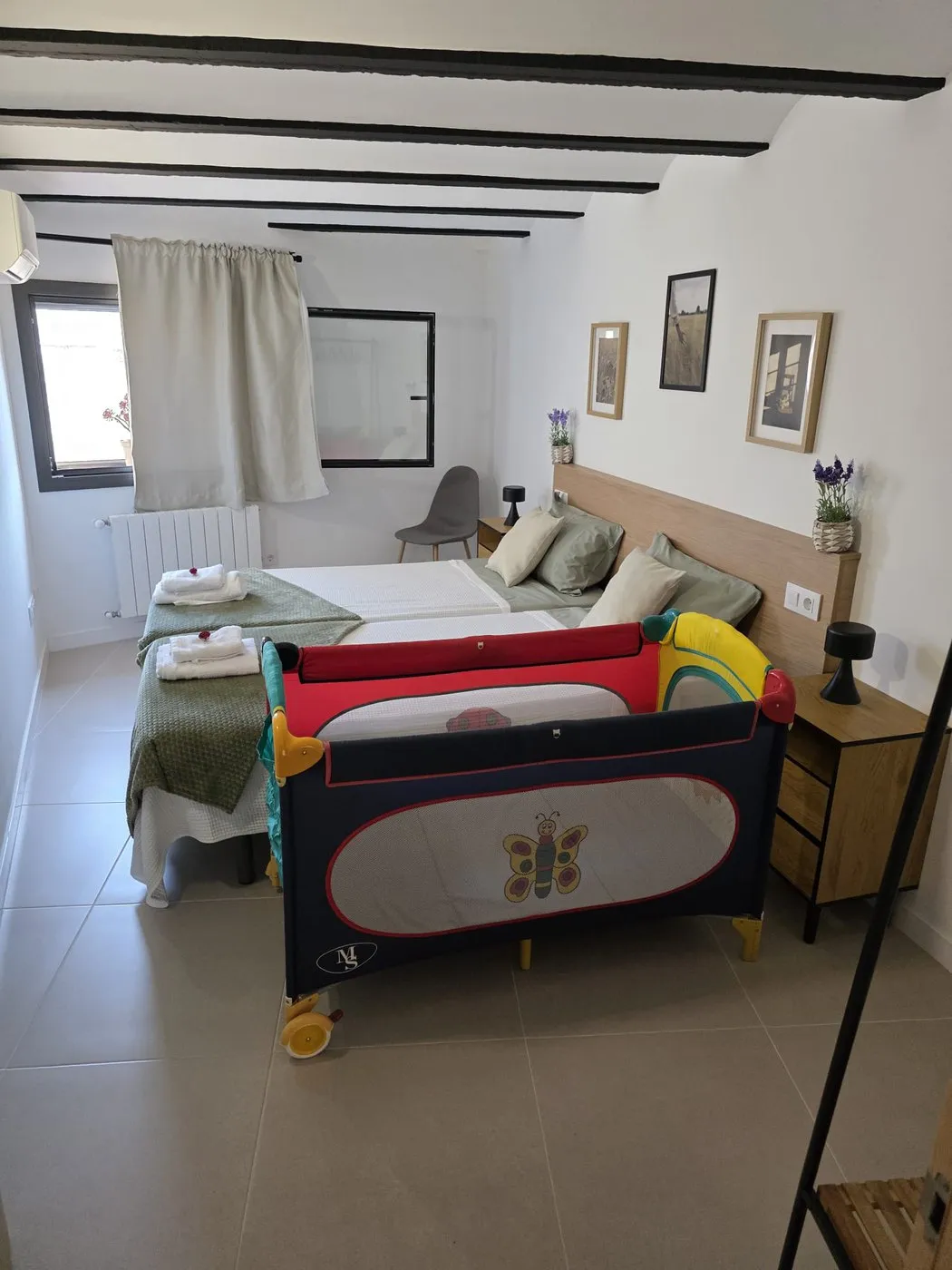 La mateixa habitació de dos llits amb un bressol de viatge muntat al costat, sota les bigues negres del sostre